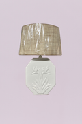 This vintage-inspired table lamp features a white ceramic base with an elegant, embossed Bird of Paradise floral motif. The base is designed in a distinctive octagonal, flat-panel shape, offering a clean and architectural silhouette. It is paired with a textured natural raffia or grasscloth drum shade, providing a beautiful contrast between the smooth glazed ceramic and the organic, tactile weave of the shade.