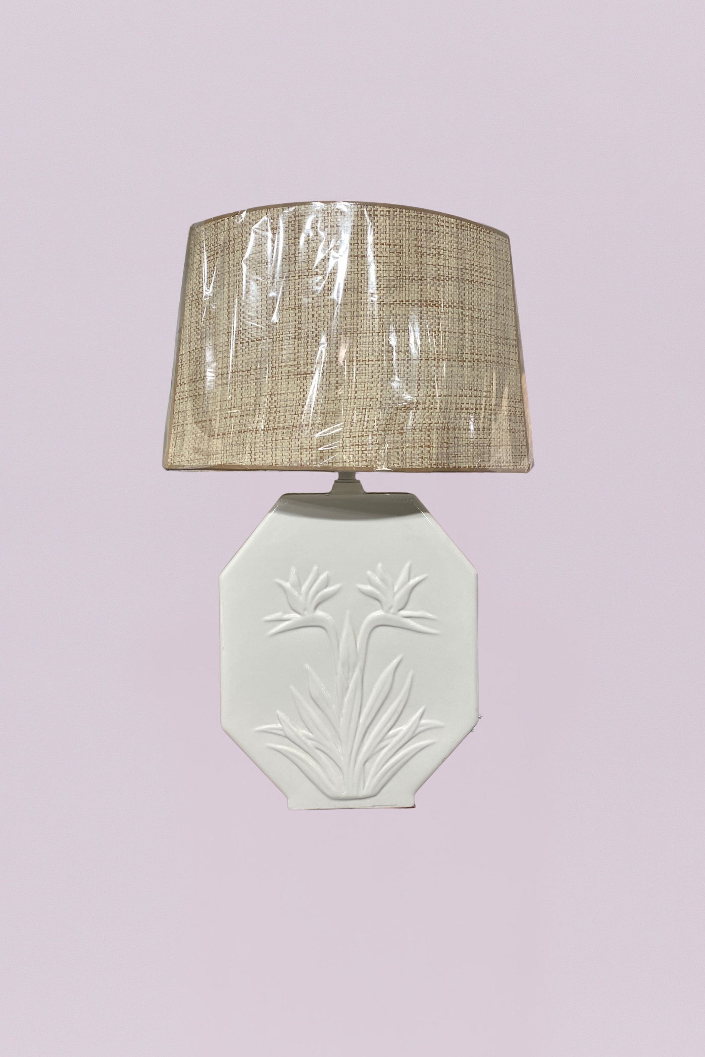 This vintage-inspired table lamp features a white ceramic base with an elegant, embossed Bird of Paradise floral motif. The base is designed in a distinctive octagonal, flat-panel shape, offering a clean and architectural silhouette. It is paired with a textured natural raffia or grasscloth drum shade, providing a beautiful contrast between the smooth glazed ceramic and the organic, tactile weave of the shade.