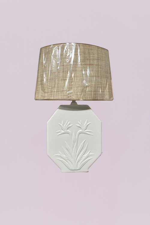 This vintage-inspired table lamp features a white ceramic base with an elegant, embossed Bird of Paradise floral motif. The base is designed in a distinctive octagonal, flat-panel shape, offering a clean and architectural silhouette. It is paired with a textured natural raffia or grasscloth drum shade, providing a beautiful contrast between the smooth glazed ceramic and the organic, tactile weave of the shade.