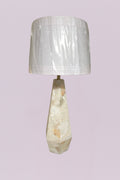 This sophisticated table lamp features a geometric, faceted base carved from natural quartz, showcasing a stunning array of organic veining in cream, white, and subtle earthy tones. The sharp, angular lines of the base provide a modern architectural feel, beautifully contrasted by the soft, clean silhouette of the white linen drum shade.