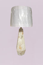 This sophisticated table lamp features a geometric, faceted base carved from natural quartz, showcasing a stunning array of organic veining in cream, white, and subtle earthy tones. The sharp, angular lines of the base provide a modern architectural feel, beautifully contrasted by the soft, clean silhouette of the white linen drum shade.