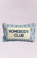 A charming, small rectangular needlepoint pillow featuring the phrase "HOMEBODY CLUB" vertically embroidered in navy blue on a cream-colored background. The pillow has a light blue border decorated with a pattern of small pink flowers and green vines, giving it a cozy, vintage, or cottage-core aesthetic. It would be a perfect decorative accent for a chair, sofa, or bed.