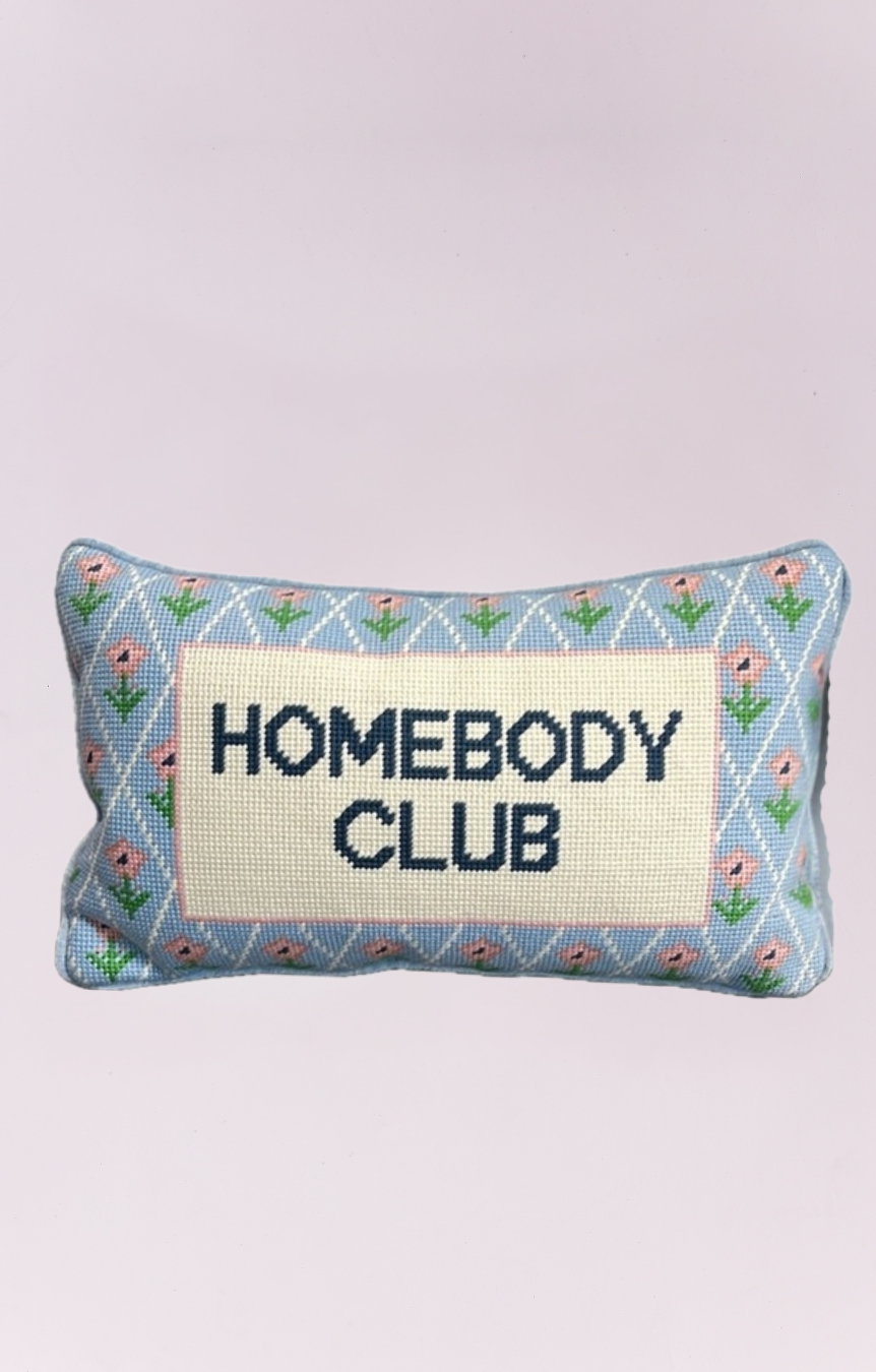A charming, small rectangular needlepoint pillow featuring the phrase "HOMEBODY CLUB" vertically embroidered in navy blue on a cream-colored background. The pillow has a light blue border decorated with a pattern of small pink flowers and green vines, giving it a cozy, vintage, or cottage-core aesthetic. It would be a perfect decorative accent for a chair, sofa, or bed.