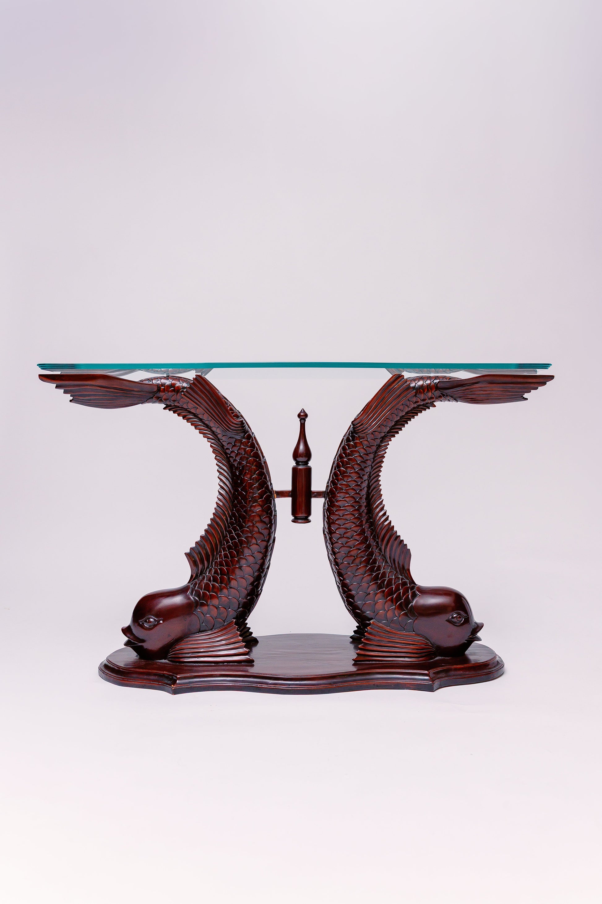 The sculptural base is formed by two carved koi fish motifs, their bodies curving gracefully upward to support a rectangular or beveled glass top. The wood (or possibly resin or composite) is richly finished to highlight details like scales, fins, and the flowing tail lines. The glass top gives it a light, open appearance and allows the artful base to be fully seen.