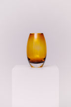 This contemporary vase features a sleek oval silhouette crafted from blown glass in a rich amber hue. The warm golden tones shift beautifully in the light, creating depth and dimension. The base is clear, adding contrast and visual grounding while showcasing the quality of the glasswork.