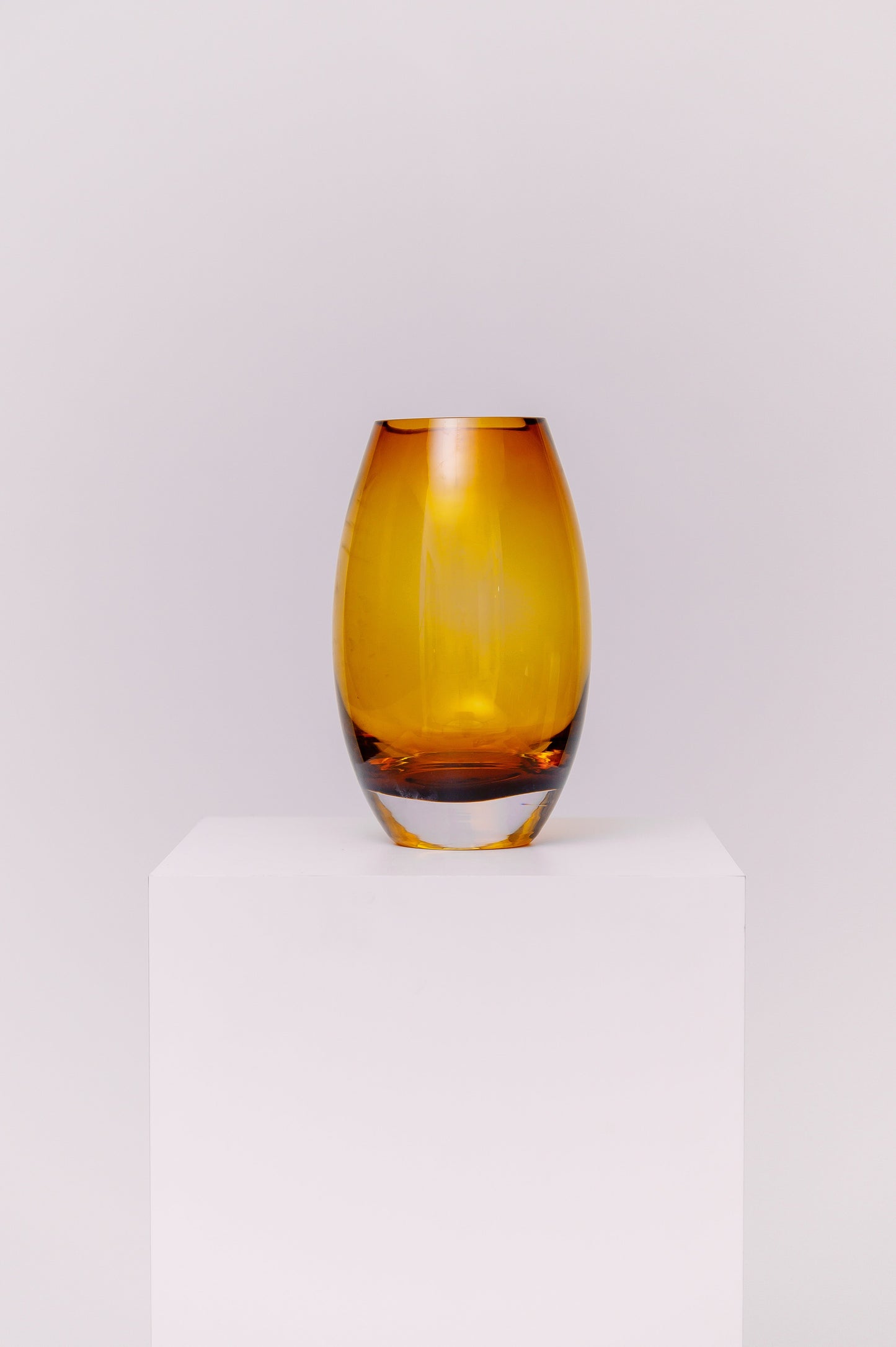 This contemporary vase features a sleek oval silhouette crafted from blown glass in a rich amber hue. The warm golden tones shift beautifully in the light, creating depth and dimension. The base is clear, adding contrast and visual grounding while showcasing the quality of the glasswork.