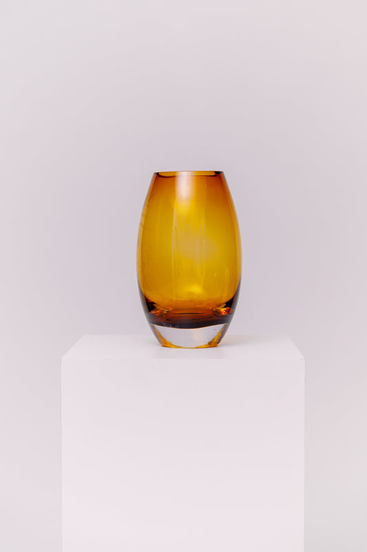 This contemporary vase features a sleek oval silhouette crafted from blown glass in a rich amber hue. The warm golden tones shift beautifully in the light, creating depth and dimension. The base is clear, adding contrast and visual grounding while showcasing the quality of the glasswork.