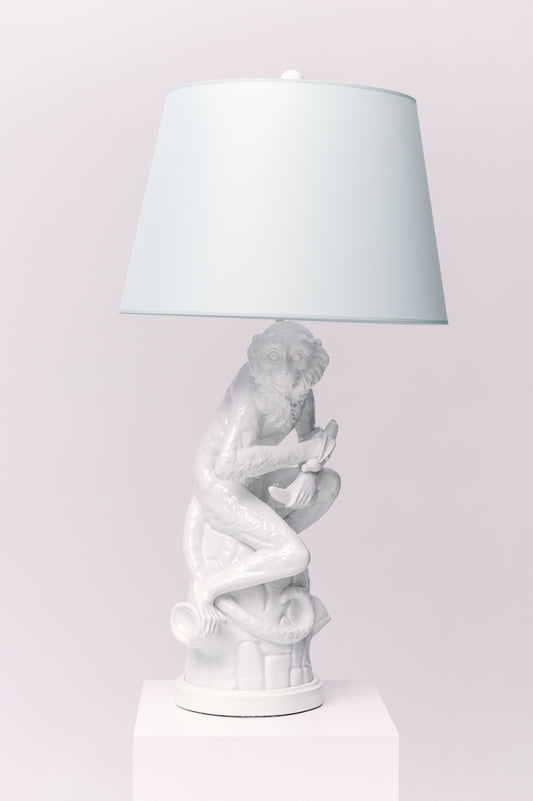 A pair of vintage Blanc de Chine monkey lamps, each with a double bulb socket. Each white porcelain monkey sits on a tree trunk holding a half eaten banana and looking curiously over his right shoulder.