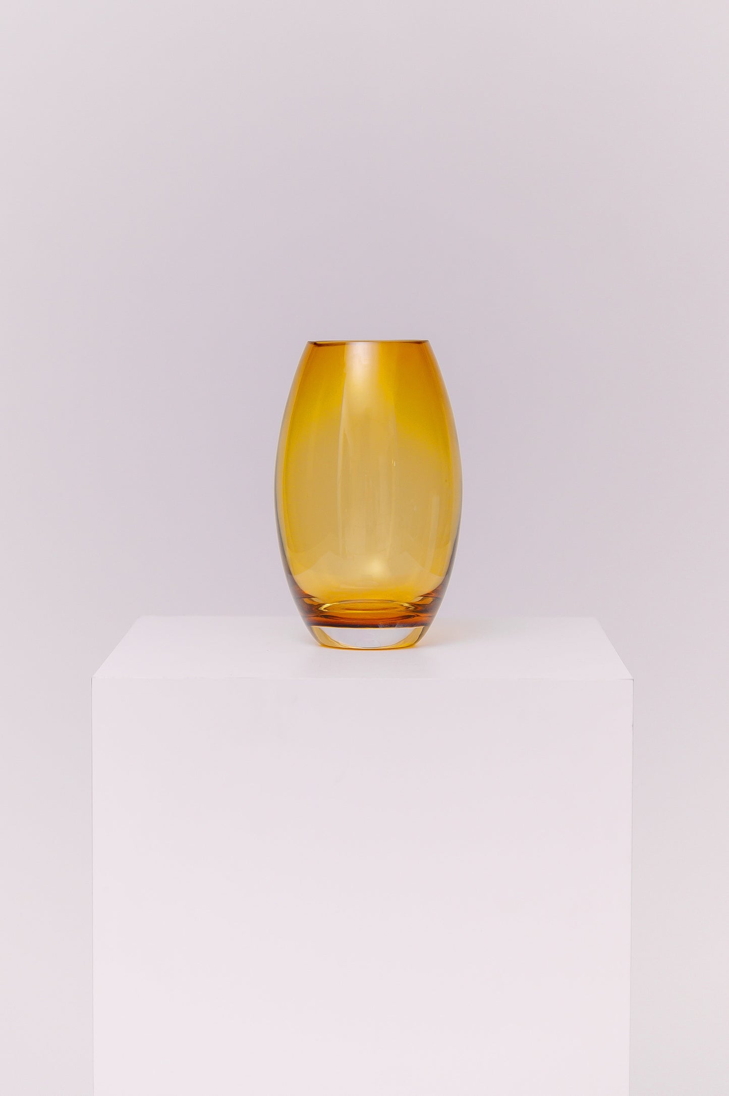 This contemporary vase features a sleek oval silhouette crafted from blown glass in a rich amber hue. The warm golden tones shift beautifully in the light, creating depth and dimension. The base is clear, adding contrast and visual grounding while showcasing the quality of the glasswork.