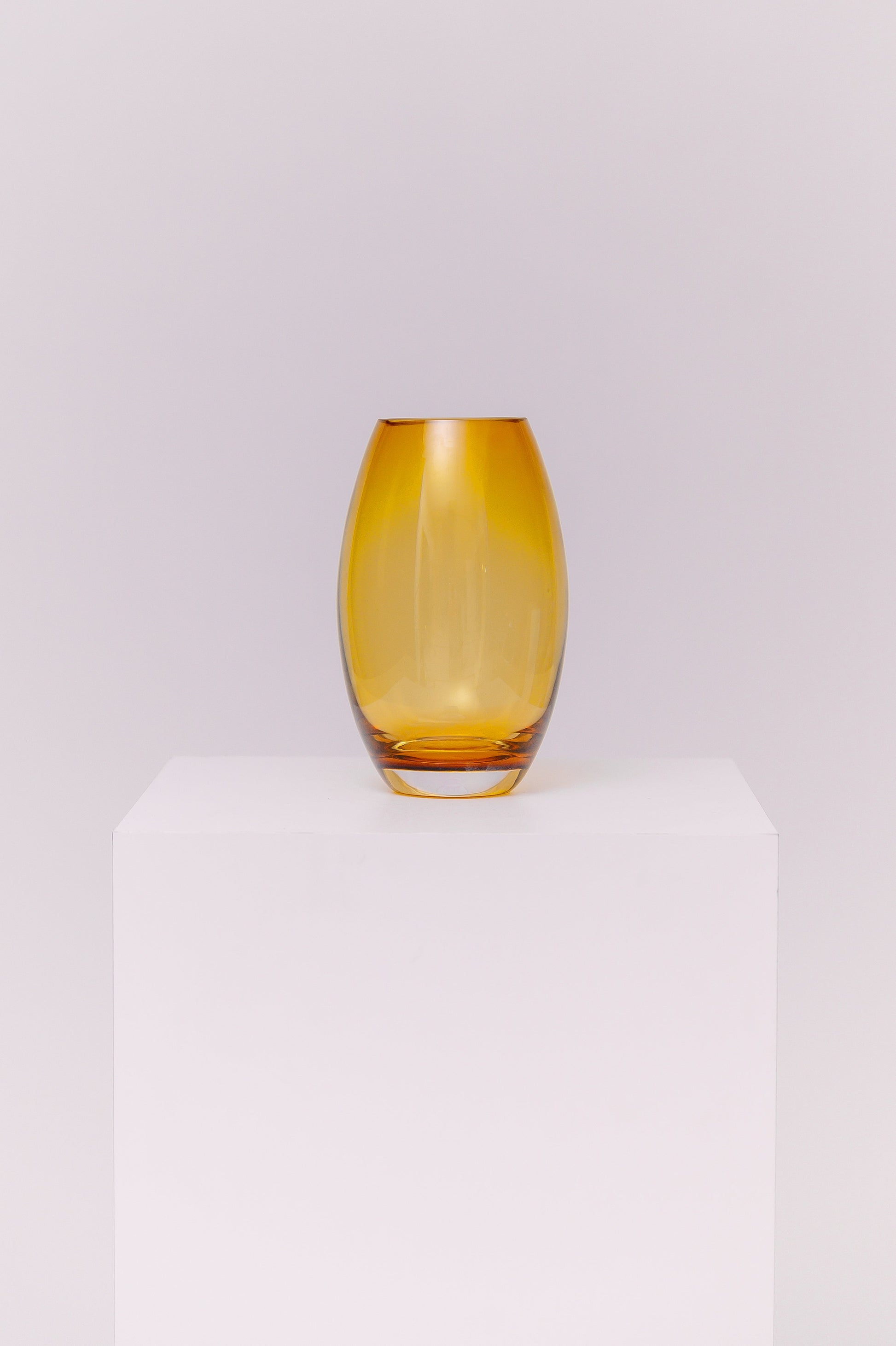 This contemporary vase features a sleek oval silhouette crafted from blown glass in a rich amber hue. The warm golden tones shift beautifully in the light, creating depth and dimension. The base is clear, adding contrast and visual grounding while showcasing the quality of the glasswork.