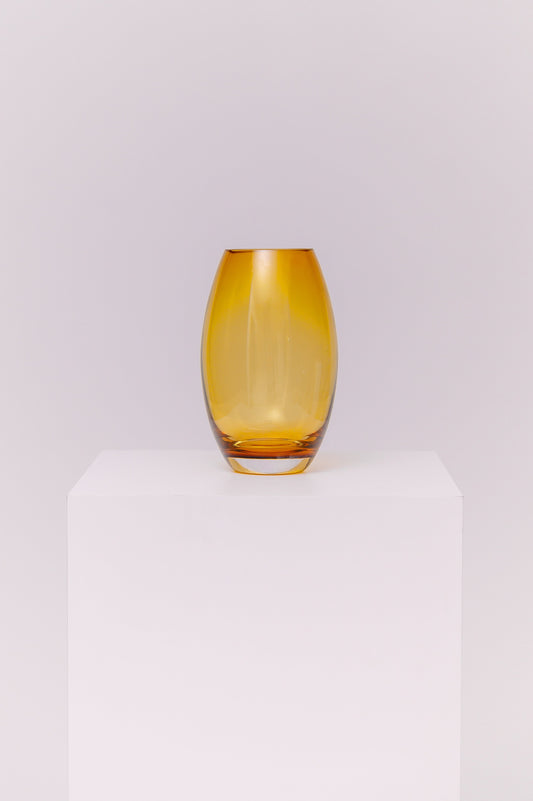 This contemporary vase features a sleek oval silhouette crafted from blown glass in a rich amber hue. The warm golden tones shift beautifully in the light, creating depth and dimension. The base is clear, adding contrast and visual grounding while showcasing the quality of the glasswork.
