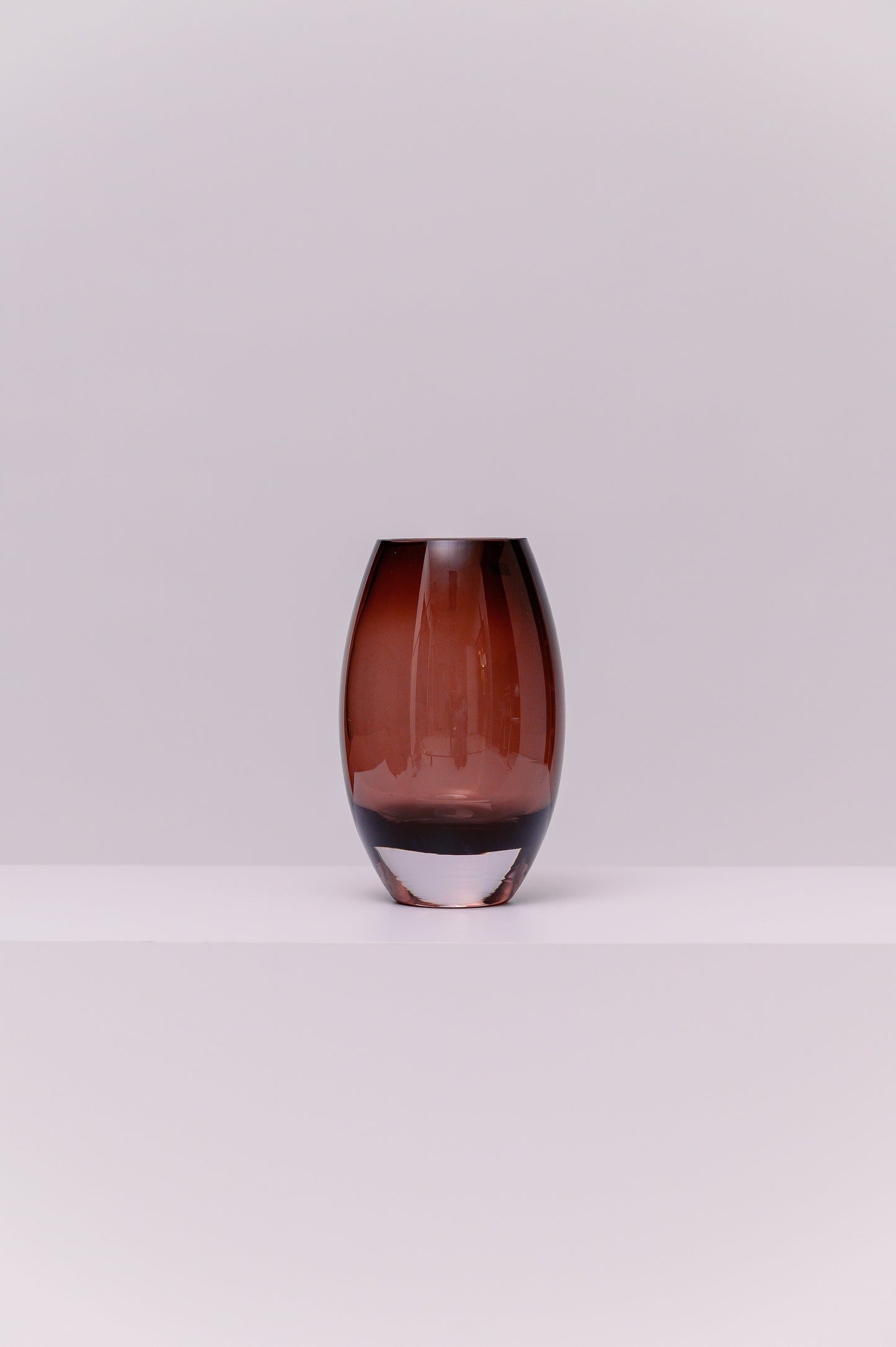 This contemporary vase features a softly rounded oval silhouette crafted from blown glass in a deep smoky purple tone. The color transitions subtly to a clear base, highlighting the thickness and quality of the glass. Its polished surface catches and reflects light, adding warmth and depth to any setting.