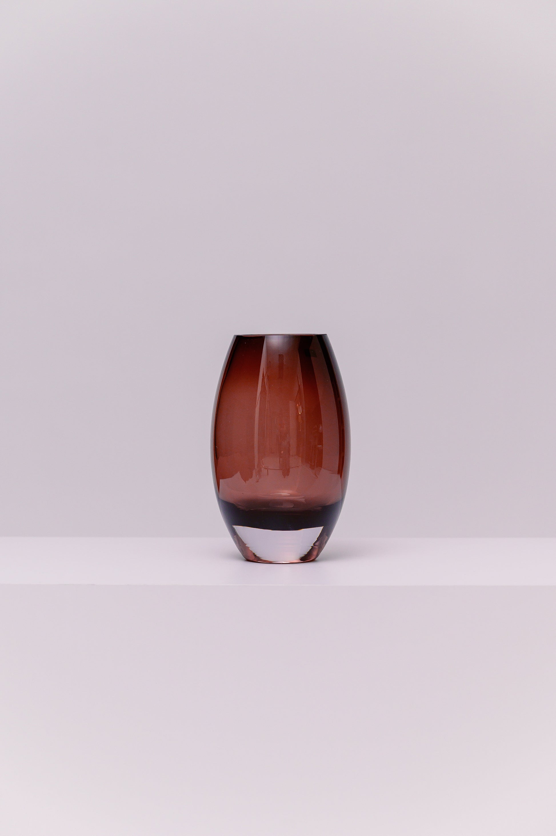 This contemporary vase features a softly rounded oval silhouette crafted from blown glass in a deep smoky purple tone. The color transitions subtly to a clear base, highlighting the thickness and quality of the glass. Its polished surface catches and reflects light, adding warmth and depth to any setting.