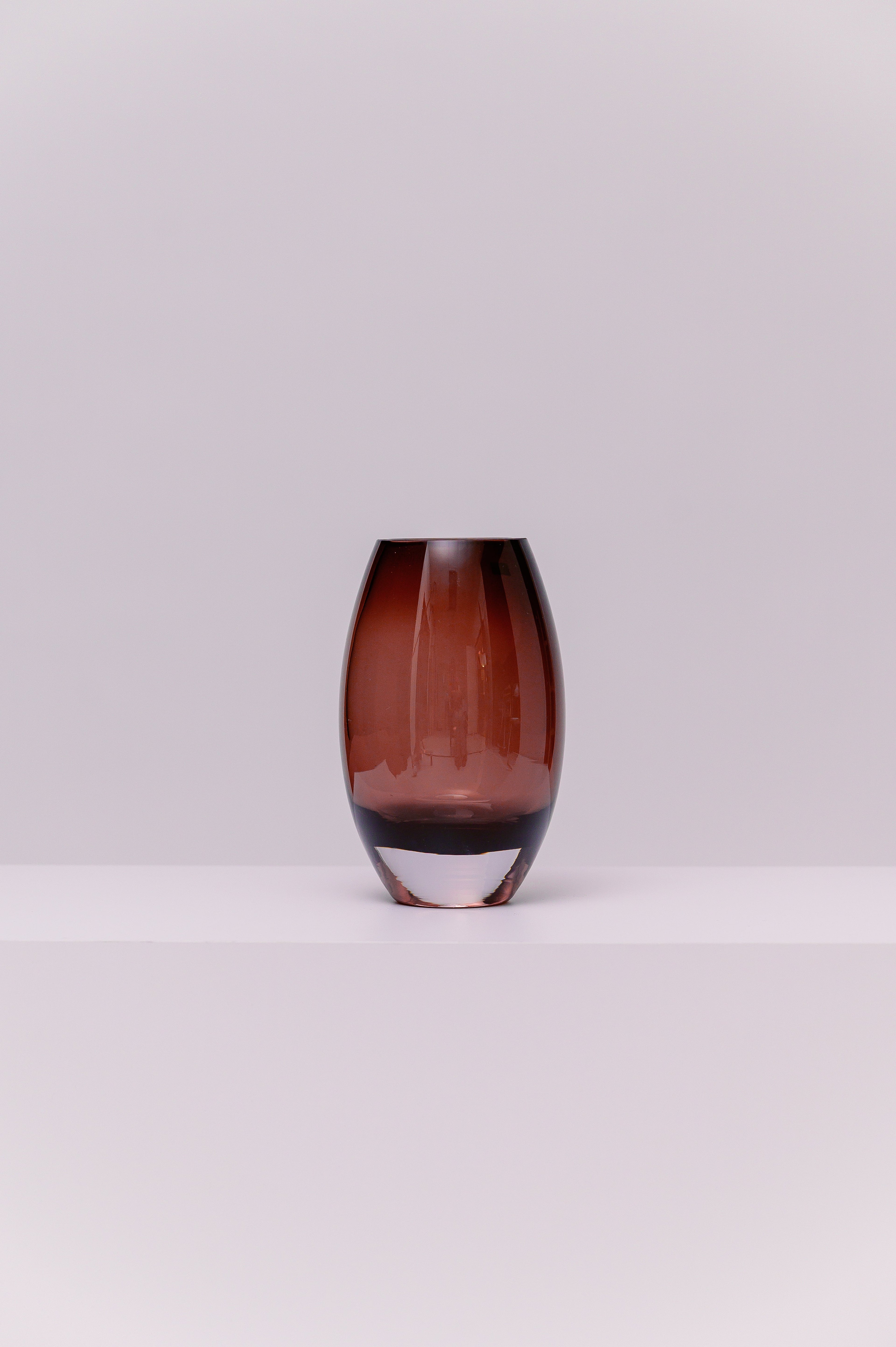 This contemporary vase features a softly rounded oval silhouette crafted from blown glass in a deep smoky purple tone. The color transitions subtly to a clear base, highlighting the thickness and quality of the glass. Its polished surface catches and reflects light, adding warmth and depth to any setting.
