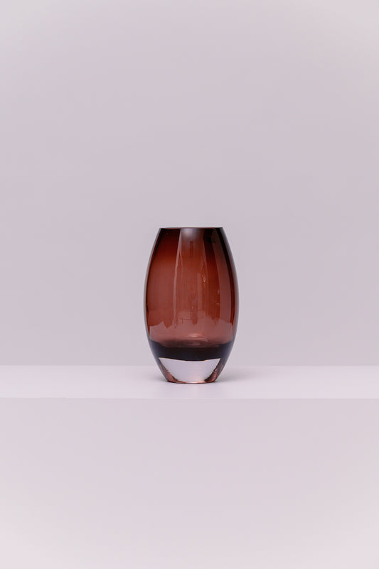 This contemporary vase features a softly rounded oval silhouette crafted from blown glass in a deep smoky purple tone. The color transitions subtly to a clear base, highlighting the thickness and quality of the glass. Its polished surface catches and reflects light, adding warmth and depth to any setting.