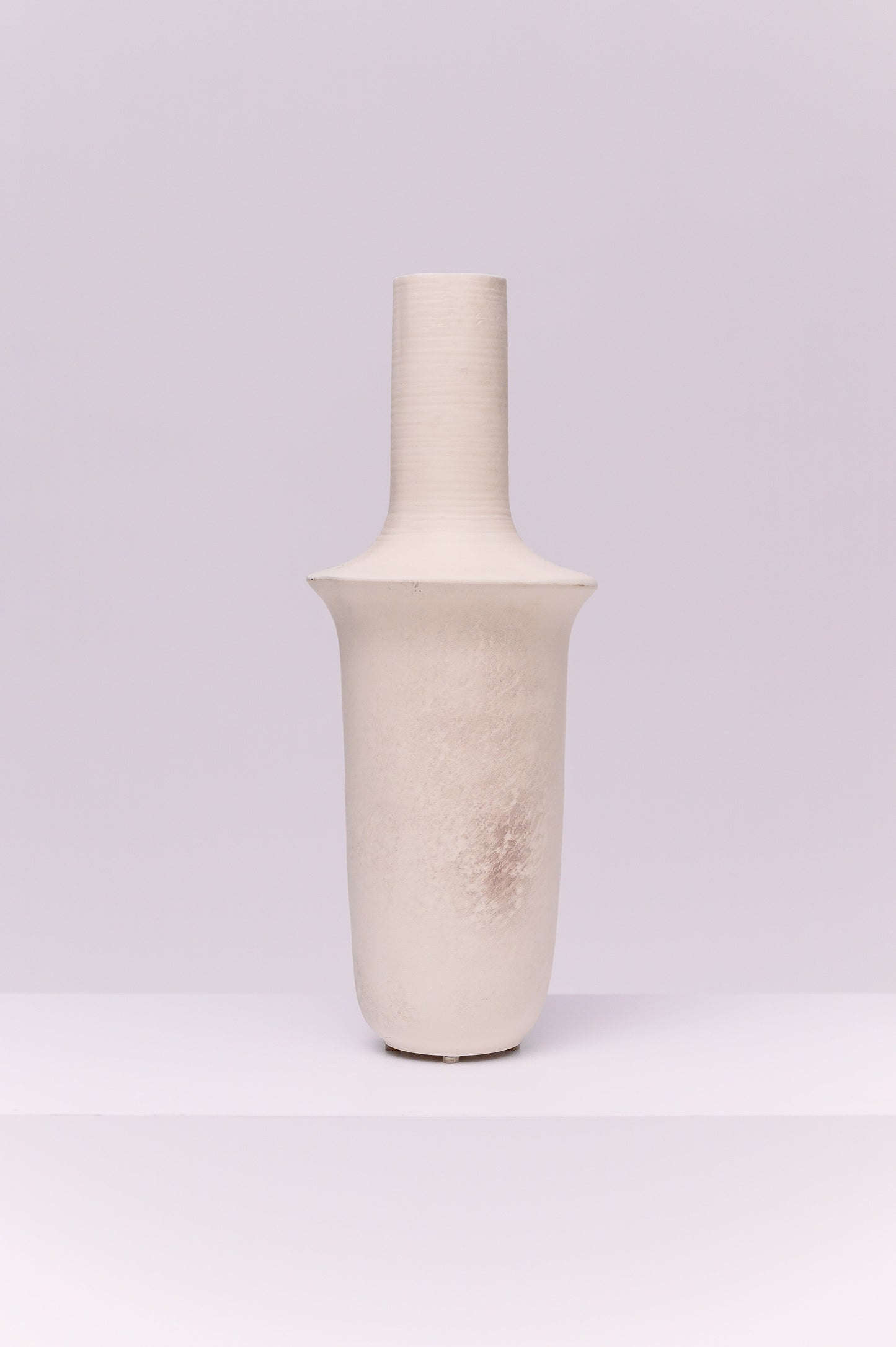 This striking vase features a tall, elongated silhouette with a cylindrical neck that flares into a broad collar before tapering to a rounded base. The matte off-white finish with subtle tonal variations gives it a raw, organic texture. Its sculptural form makes it a modern statement piece, ideal for display on its own or styled with tall branches or dried botanicals.