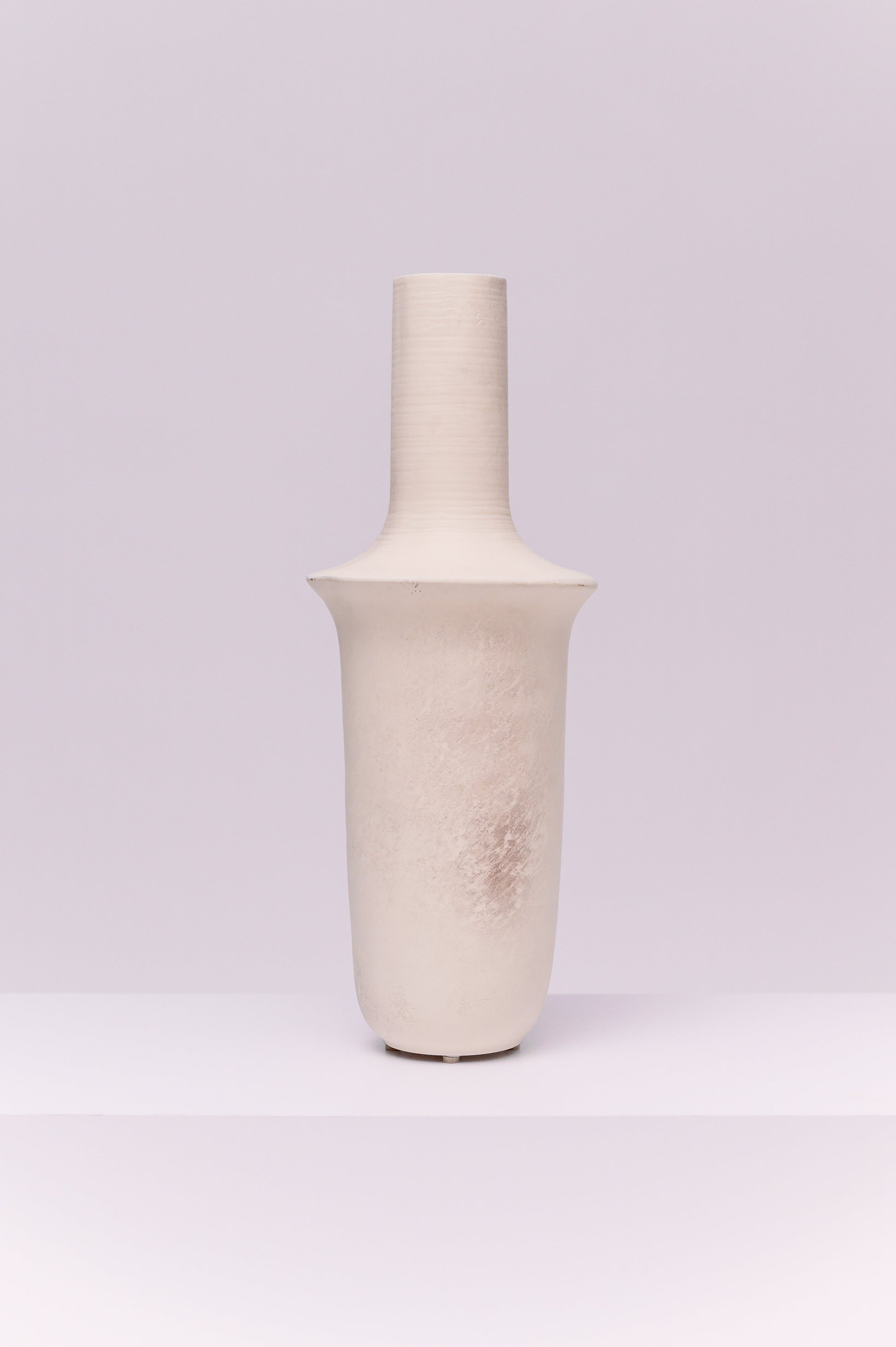 This striking vase features a tall, elongated silhouette with a cylindrical neck that flares into a broad collar before tapering to a rounded base. The matte off-white finish with subtle tonal variations gives it a raw, organic texture. Its sculptural form makes it a modern statement piece, ideal for display on its own or styled with tall branches or dried botanicals.