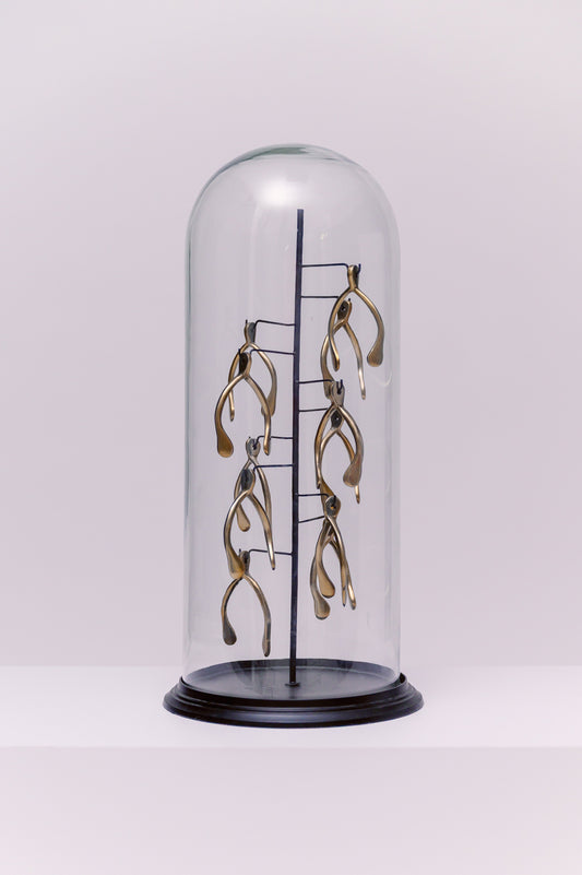This decorative piece features a collection of golden wishbones.  The entire sculpture is housed within a clear glass cloche, or bell jar, which sits on a black circular base. The wishbones are mounted at different levels and angles, creating a whimsical and artistic display. The use of a cloche highlights the wishbones as a central feature, transforming them from simple objects into a curated work of art.