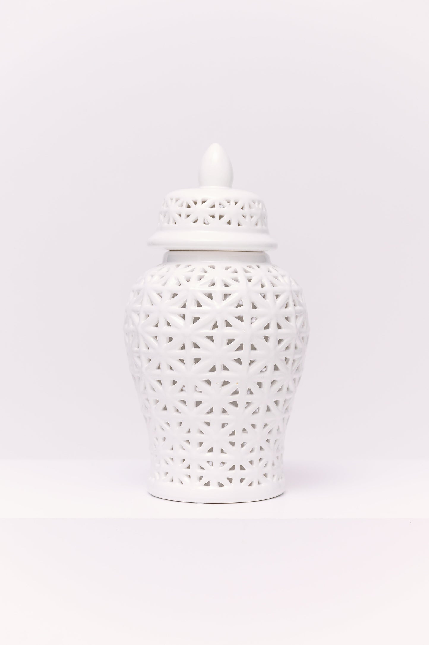 This elegant white porcelain ginger jar features a reticulated (pierced) geometric lattice design, giving it a delicate, airy quality. The pattern, formed by interlocking star-like shapes, allows light to filter through, creating subtle shadows and dimension. Finished with a matching pierced lid and pointed finial, the jar is a timeless decorative accent with a clean, modern chinoiserie appeal.