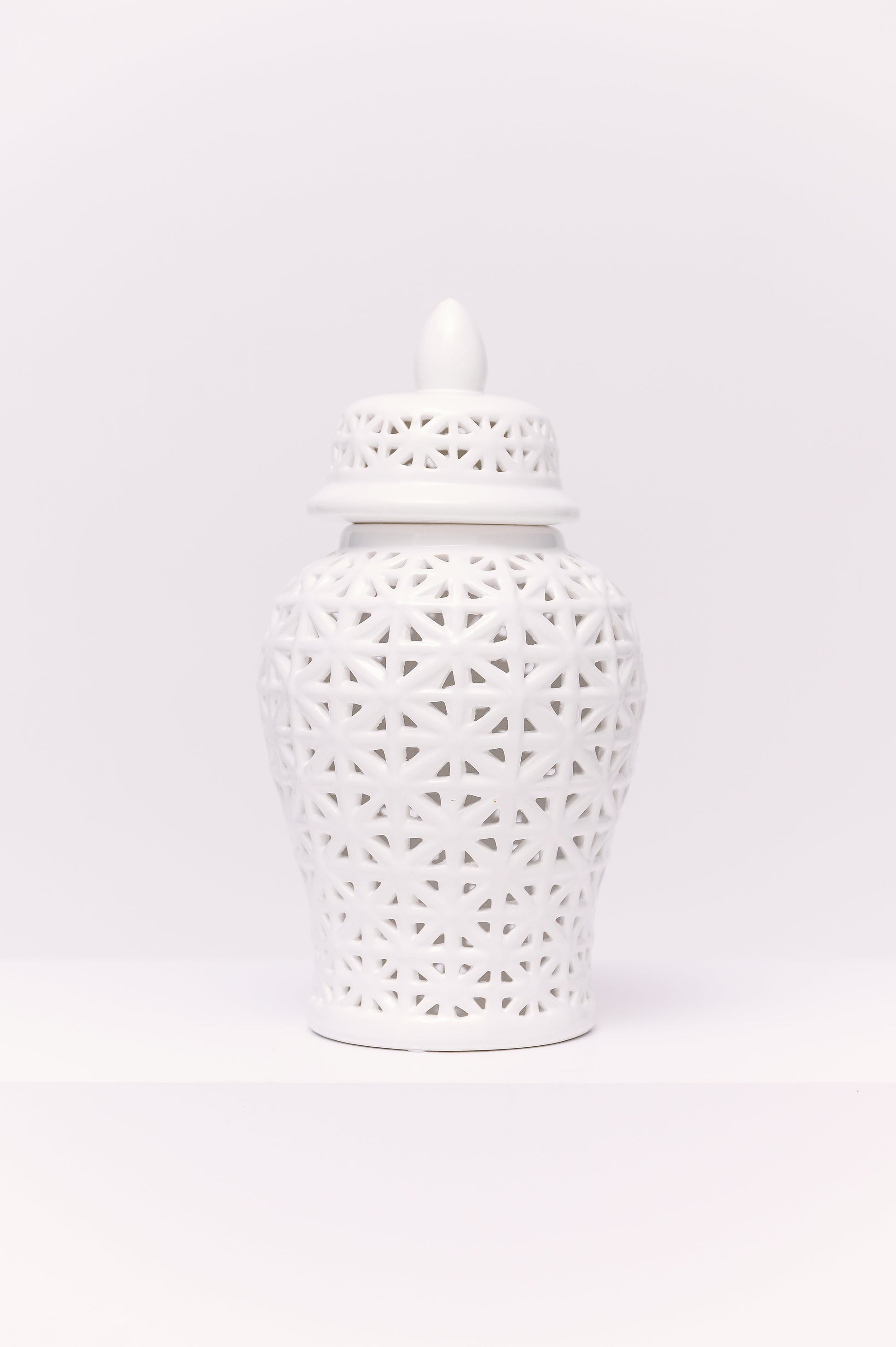 This elegant white porcelain ginger jar features a reticulated (pierced) geometric lattice design, giving it a delicate, airy quality. The pattern, formed by interlocking star-like shapes, allows light to filter through, creating subtle shadows and dimension. Finished with a matching pierced lid and pointed finial, the jar is a timeless decorative accent with a clean, modern chinoiserie appeal.